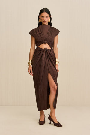 Woman wearing a brown structured midi dress crafted from plisse fabric that features a mock neck, front slit, and a knotted cutout detail below the bust, paired with brown heels. 