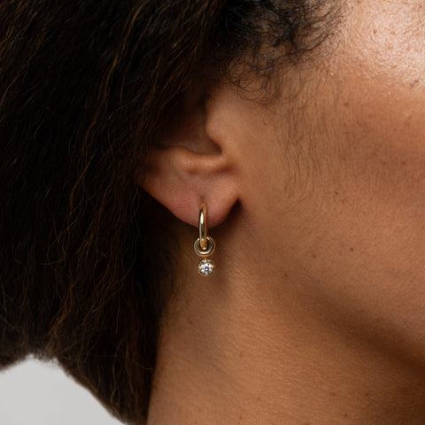 Woman wearing an 18k yellow gold mini hoop earring that features three 18k yellow gold annulets, one with a round white diamond charm. 