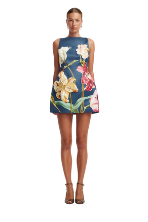 A model wearing a sleeveless navy blue shift dress with a large, colorful floral print of lilies and tulips, paired with brown strappy high-heeled sandals against a plain white background.
