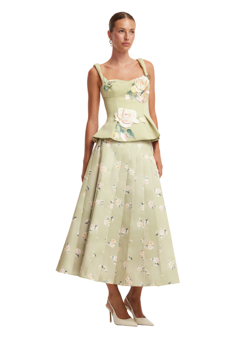 A model wearing a light sage green sleeveless midi dress with a floral print, featuring a structured peplum bodice with large 3D rose appliques and a matching pleated skirt, paired with beige high heels against a white background.