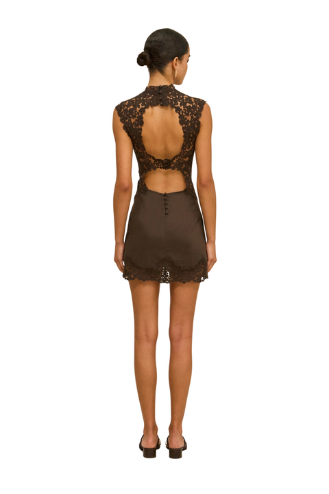 A woman viewed from the back wearing a sleeveless brown mini dress with a lace open back and a small waist cutout, paired with black heels, against a plain white background.
