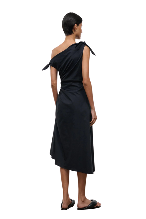 A woman shown from the back wearing a black one-shoulder midi dress with a tie detail on the shoulder, paired with black slide sandals, against a plain white background.