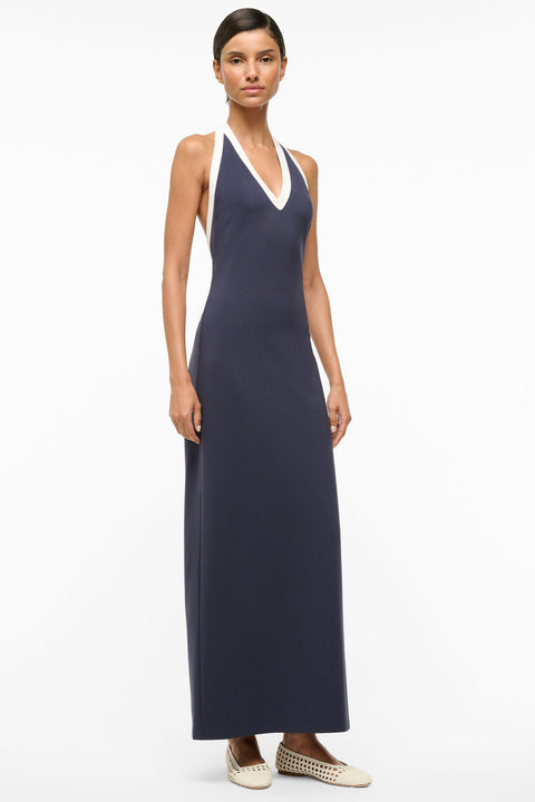 Side angle of a woman wearing a navy maxi dress with a halter neckline that goes into a deep-v and low cut back, trimmed in white. 