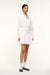 Side view of a woman wearing a long sleeve, white mini dress that features a collar and button down detail and a curved yoke at the hem. 
