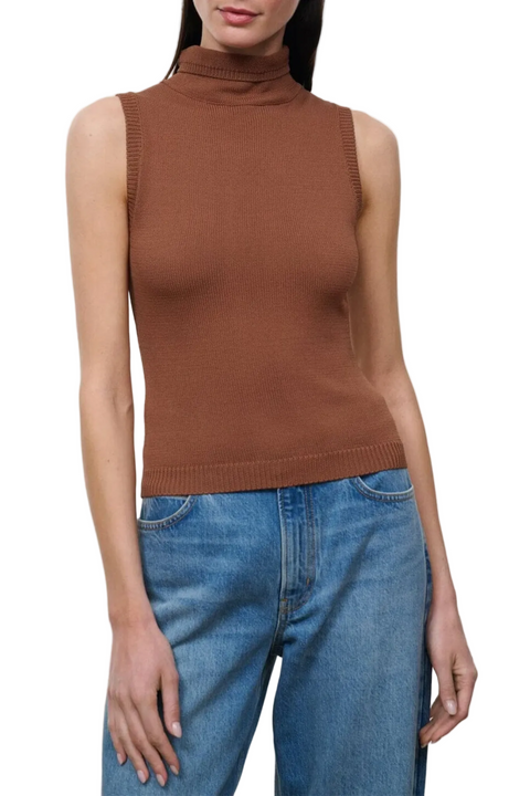 A woman wearing a sleeveless ribbed mock neck top in a warm rust brown color, paired with blue jeans, against a plain light gray studio background.