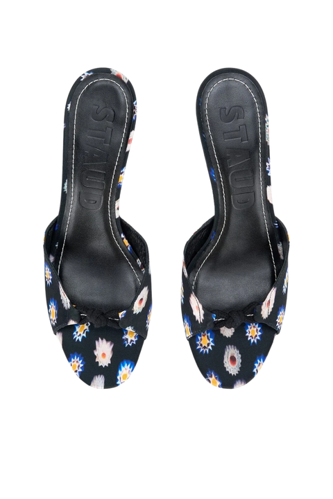 A pair of black slide sandals with a colorful floral print, featuring a knot detail on the toe strap and the brand name 'STAUD' embossed on the insole, shown against a plain white background.