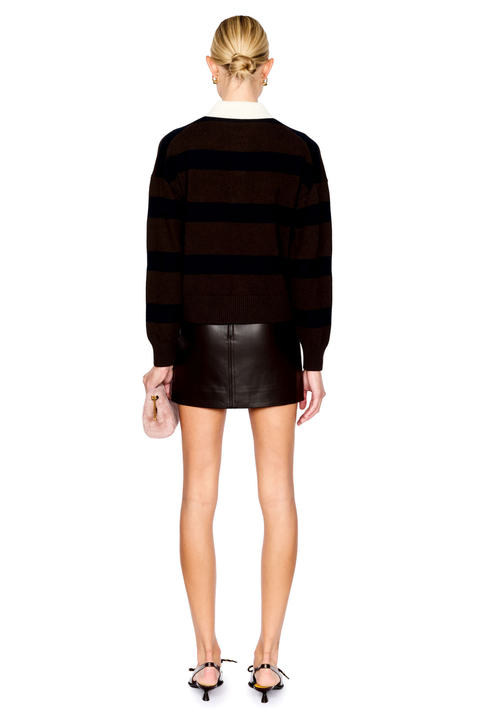 A model shown from the back wearing a dark brown and navy horizontally striped knit sweater, a brown leather mini skirt, and brown strappy heels, holding a small pink clutch against a plain white background.