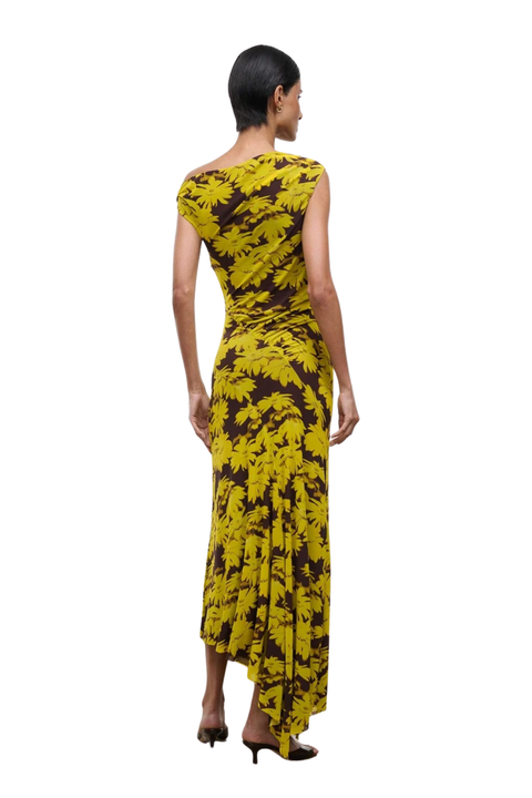 A woman shown from the back wearing a sleeveless, floor-length dress with a bold yellow and dark brown abstract floral pattern, photographed against a plain white studio background.