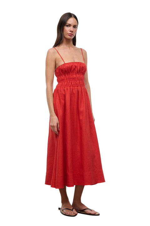 A woman modeling a bright red midi dress with thin spaghetti straps, a smocked bodice, and a flowing skirt, paired with brown sandals against a plain white background.