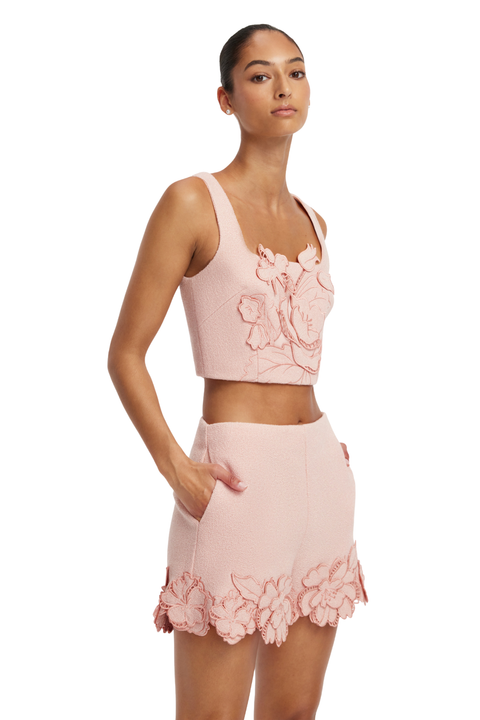 A model wearing a light pink two-piece set consisting of a sleeveless crop top and a matching high-waisted mini skirt, both featuring 3D floral applique details.