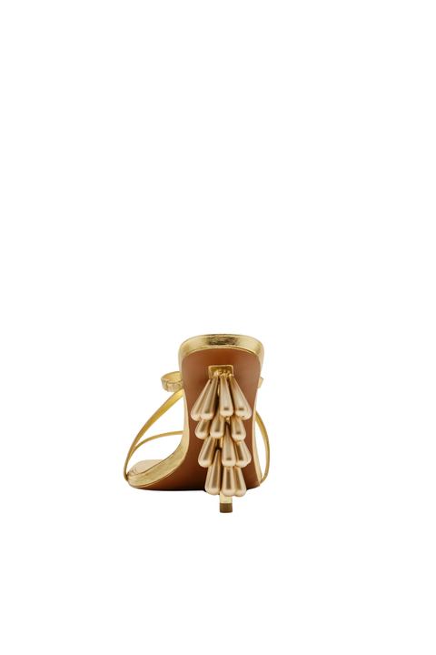 A single women's heeled sandal shown from the back on a plain white background. The sandal has a tan leather upper with a gold metallic heel, toe cap, and decorative hanging fringe detail.