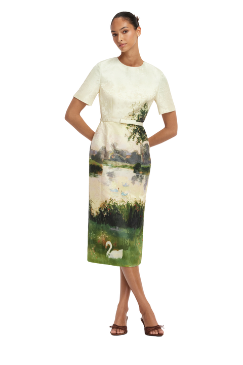 A woman modeling a short-sleeve midi dress with a cream top and a scenic landscape print on the skirt, featuring green fields, trees, water, and a white swan, set against a plain white background.