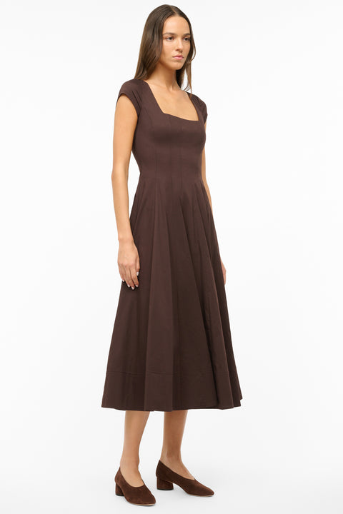Side view of a woman wearing a brown midi dress that features cap sleeves, a square neck, corset styles paneling, and an A-line skirt, paired with brown kitten heels. 