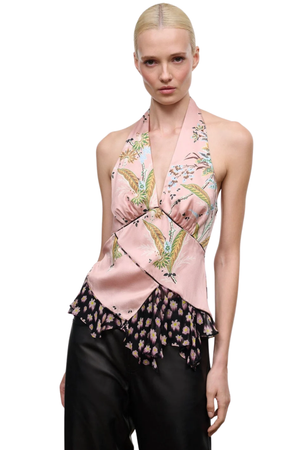 A model wearing a halter-neck top in light pink silk with a green and gold bird print, paired with black pants, against a plain white background.