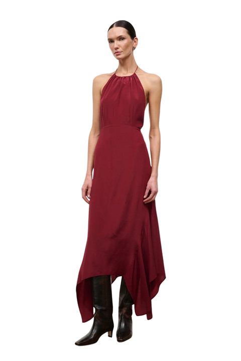 A woman modeling a sleeveless halter-neck maxi dress in deep burgundy with an asymmetrical draped hem, paired with black knee-high boots, against a plain white background.