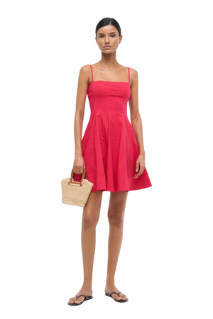 A woman modeling a bright cherry sleeveless mini dress with thin straps and a flared skirt, holding a small woven straw handbag against a plain white background.