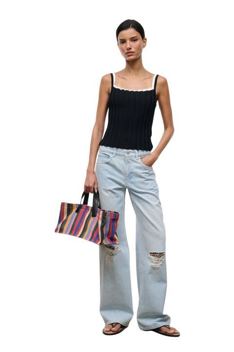 A model stands against a white background wearing a black ribbed camisole, light-wash distressed wide-leg jeans, and sandals, while holding a colorful striped tote bag.