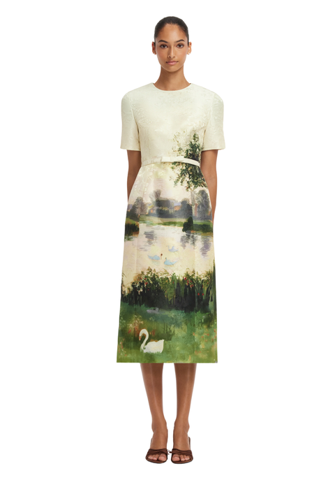 A model wearing a short-sleeve midi dress with a cream-colored textured bodice and a skirt featuring a scenic landscape print with green grass, a lake, trees, and a white swan, set against a plain white background.