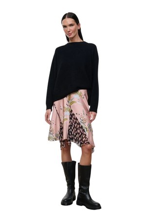 A model stands against a white background wearing a black long-sleeve knit top, a pink patterned asymmetrical skirt, and black knee-high boots.