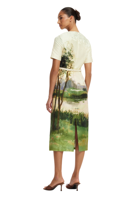 A woman shown from the back wearing a short-sleeve midi dress with a cream base and a green landscape painting print, paired with brown heeled sandals against a plain white background.