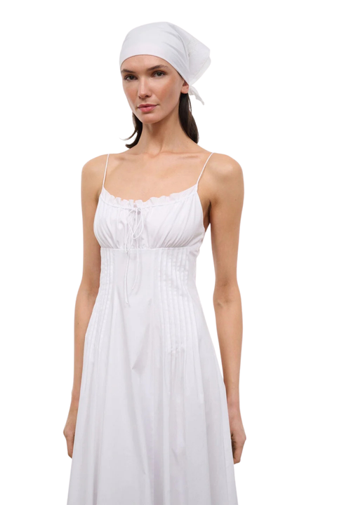 A woman modeling a white sleeveless maxi dress with thin straps, a gathered bust, and a matching white headscarf, photographed against a plain white studio background.