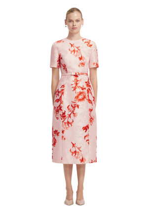 A woman modeling a short-sleeved midi dress with a red floral pattern on a light pink textured fabric, set against a plain white background.