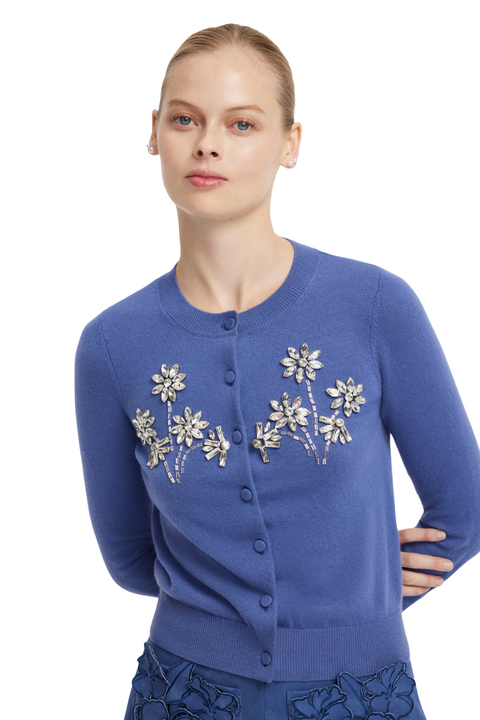 A woman modeling a blue button-front cardigan with embroidered floral details on the chest, photographed against a plain white background.