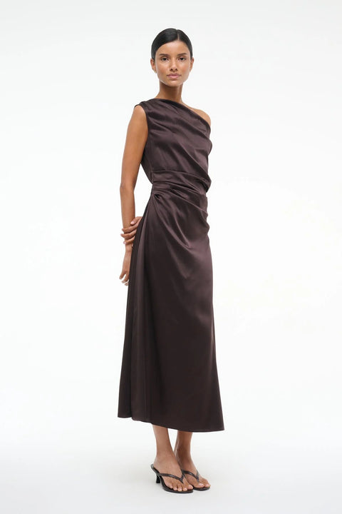 Side angle of a woman wearing an asymmetrical brown silk midi dress with an off the shoulder design and ruched draping throughout, paired with brown heels.