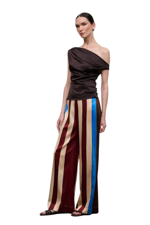 A model wearing a dark brown draped off-the-shoulder top and wide-leg trousers with vertical stripes in burgundy, beige, and blue, standing against a plain white background.