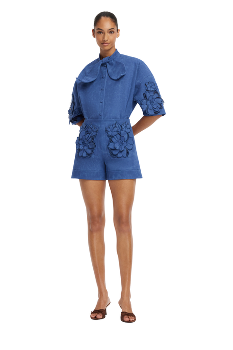 A model wearing a blue short-sleeve matching set with 3D floral applique details on the pockets and sleeves, paired with brown flat sandals, shot against a solid white background.