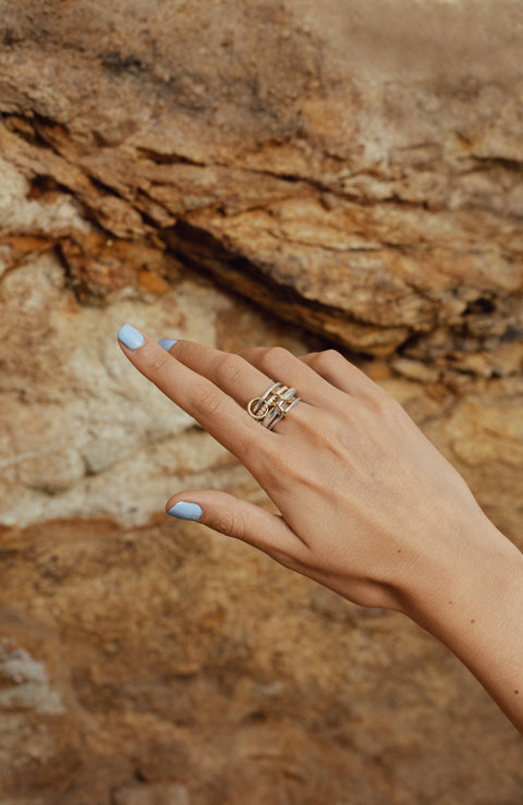 A woman's hand wearing a Spinelli Kilcollin ring that features a thin sterling silver band, a thin 18k yellow gold band with pave diamonds, a very thick sterling silver band, and another thin sterling silver band, all connected by three 18k yellow gold connector rings on her index finger.