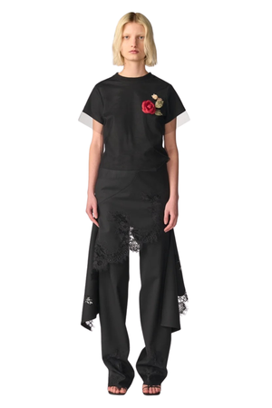 A model wearing a black short-sleeve t-shirt with a red rose graphic on the chest, paired with black wide-leg pants featuring a lace-trimmed asymmetrical hem, shot against a solid white background.