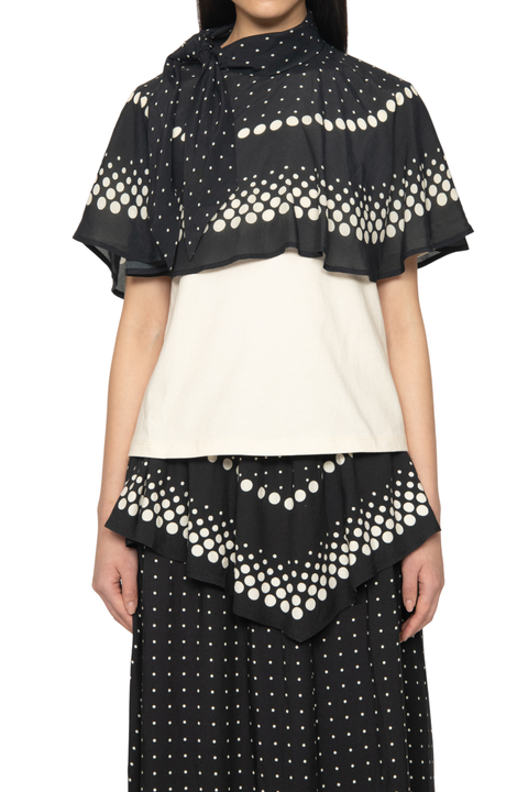 A model wearing a black and off-white color-blocked top with a draped, ruffled overlay featuring a cream polka-dot pattern, paired with matching polka-dot skirt.