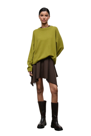 A model stands against a white background wearing an oversized olive green knit sweater, a dark brown asymmetrical mini skirt, and black leather boots.