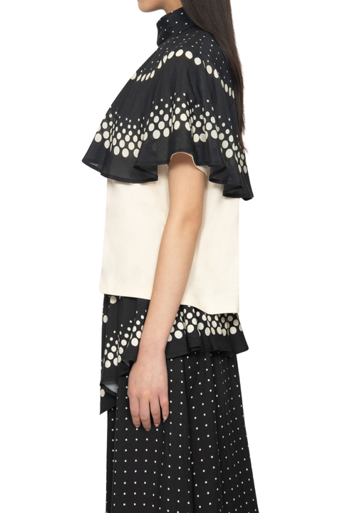 A model shown from the side wearing a black and off-white color-blocked top with dramatic ruffled sleeves and a matching black skirt featuring a white polka-dot pattern, set against a plain white background.