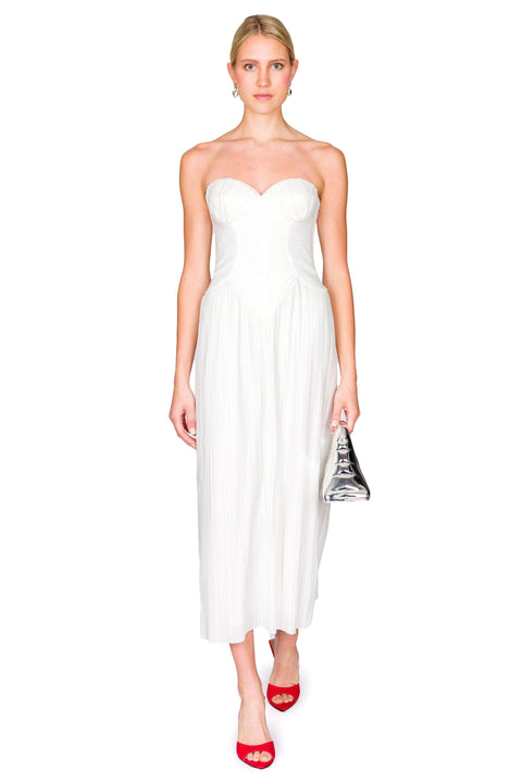 Woman wearing a white, strapless midi dress crafted from plisse fabric that features a sweetheart neckline and corset detailing, paired with red heels and a silver handbag. 