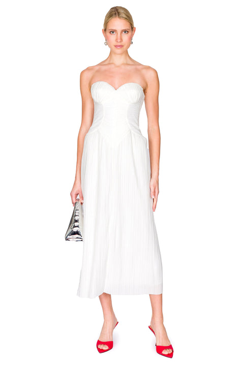 Woman wearing a white, strapless midi dress crafted from plisse fabric that features a sweetheart neckline and corset detailing, paired with red heels and a silver handbag. 