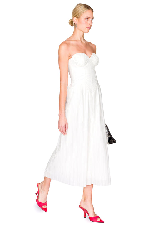 Side view of a woman wearing a white, strapless midi dress crafted from plisse fabric that features a sweetheart neckline and corset detailing, paired with red heels and a silver handbag. 