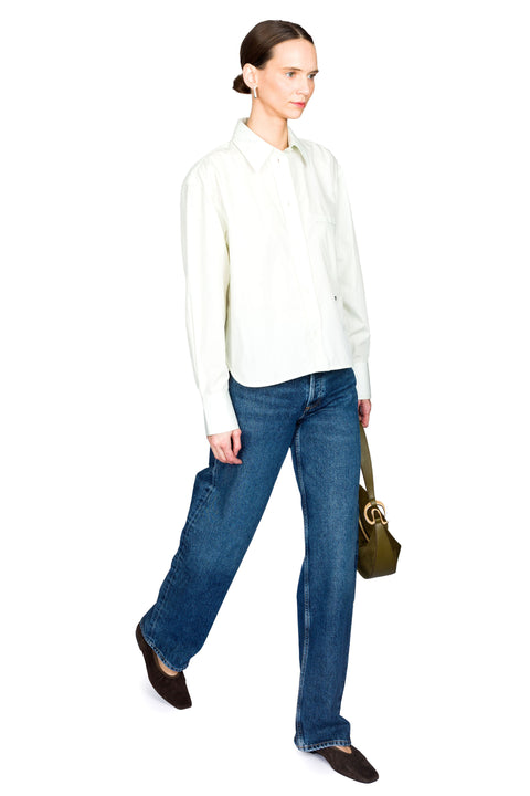 Woman taking a step wearing a light green poplin shirt and blue jeans on a white background