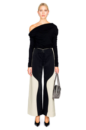 Woman wearing a black long sleeve off-shoulder top and black and cream pants on a white background