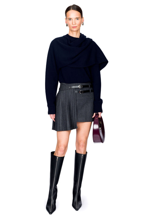 Woman wearing a navy blue draped sweater, gray pleated mini skirt, red handbag and black knee-high boots on a white background