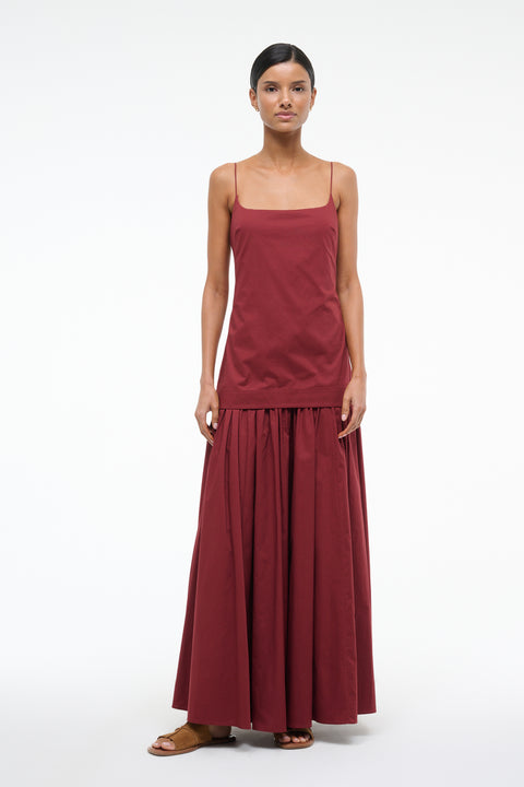Woman wearing a deep red maxi dress that features a column bodice layered over a full skirt, paired with brown sandals. 