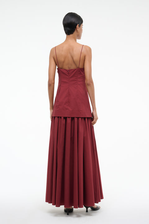 Back-facing woman wearing a deep red maxi dress that features a column bodice layered over a full skirt, paired with black kitten heels. 