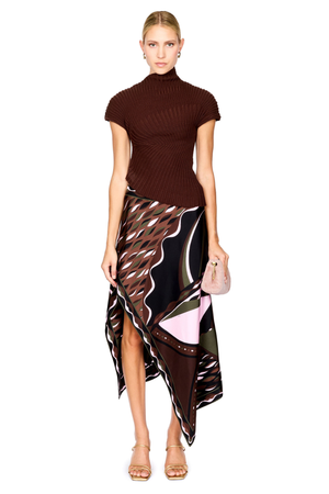 A model stands against a white studio background wearing a dark brown ribbed short-sleeve mock-neck top and a high-low wrap skirt with a black, brown, beige, and pink abstract print. She is holding a small blush pink handbag and wearing gold high-heeled sandals.