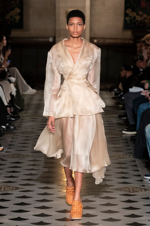 Model walking down a runway in the fitted organdy jacket with back cut-out in beige.