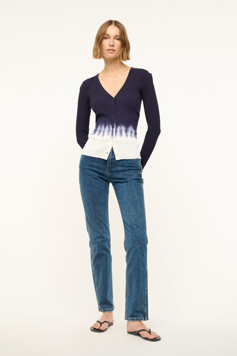 Woman wearing a fitted, rib-knit cardigan that features a deep-V and a navy to white tie dye effect, paired with jeans and sandals.  