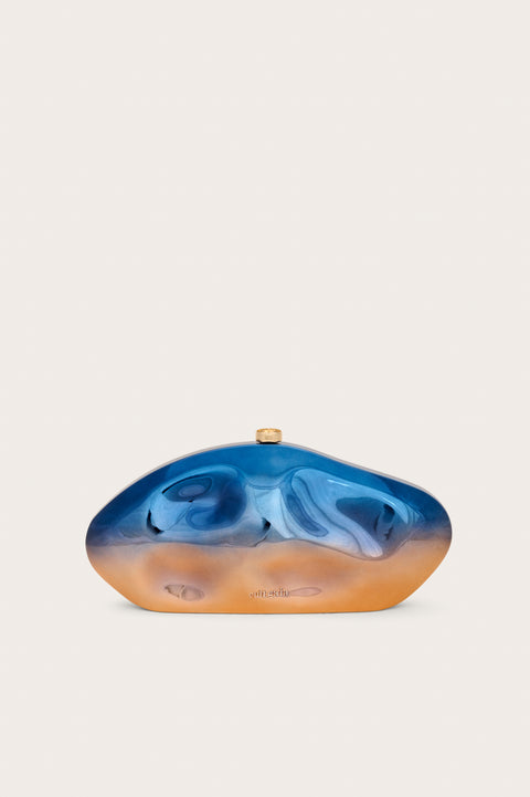 An abstract oval shaped acrylic clutch that features formed dents and a metallic blue to orange ombre effect, finished with gold hardware. 