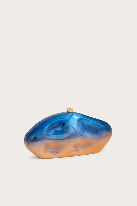 Side view of an abstract oval shaped acrylic clutch that features formed dents and a metallic blue to orange ombre effect, finished with gold hardware. 