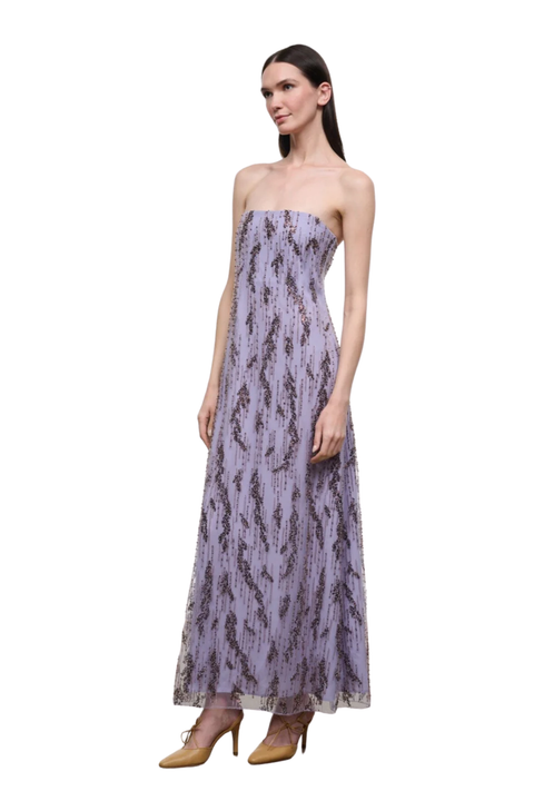 A woman modeling a strapless lavender maxi dress with a dark abstract print, paired with tan high heels, set against a plain white background.