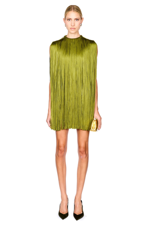 A model stands against a white background wearing a sleeveless olive green mini dress with a high mock neck, paired with dark green pointed-toe heels and holding a small gold clutch.